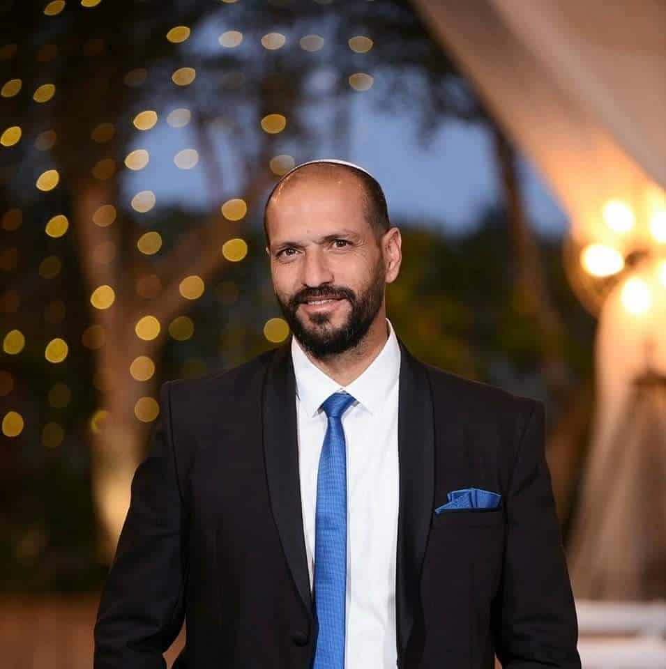 גבר עם זקן וחליפה כהה ועניבה כחולה מחייך לעבר המצלמה ברקע מואר.