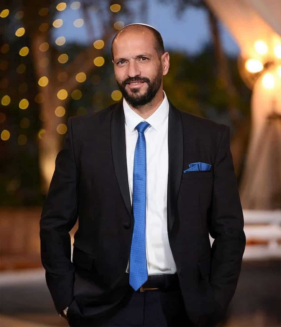 גבר עם כיפה לבנה לבוש בחליפה שחורה ועניבה כחולה עומד ברקע מטושטש.